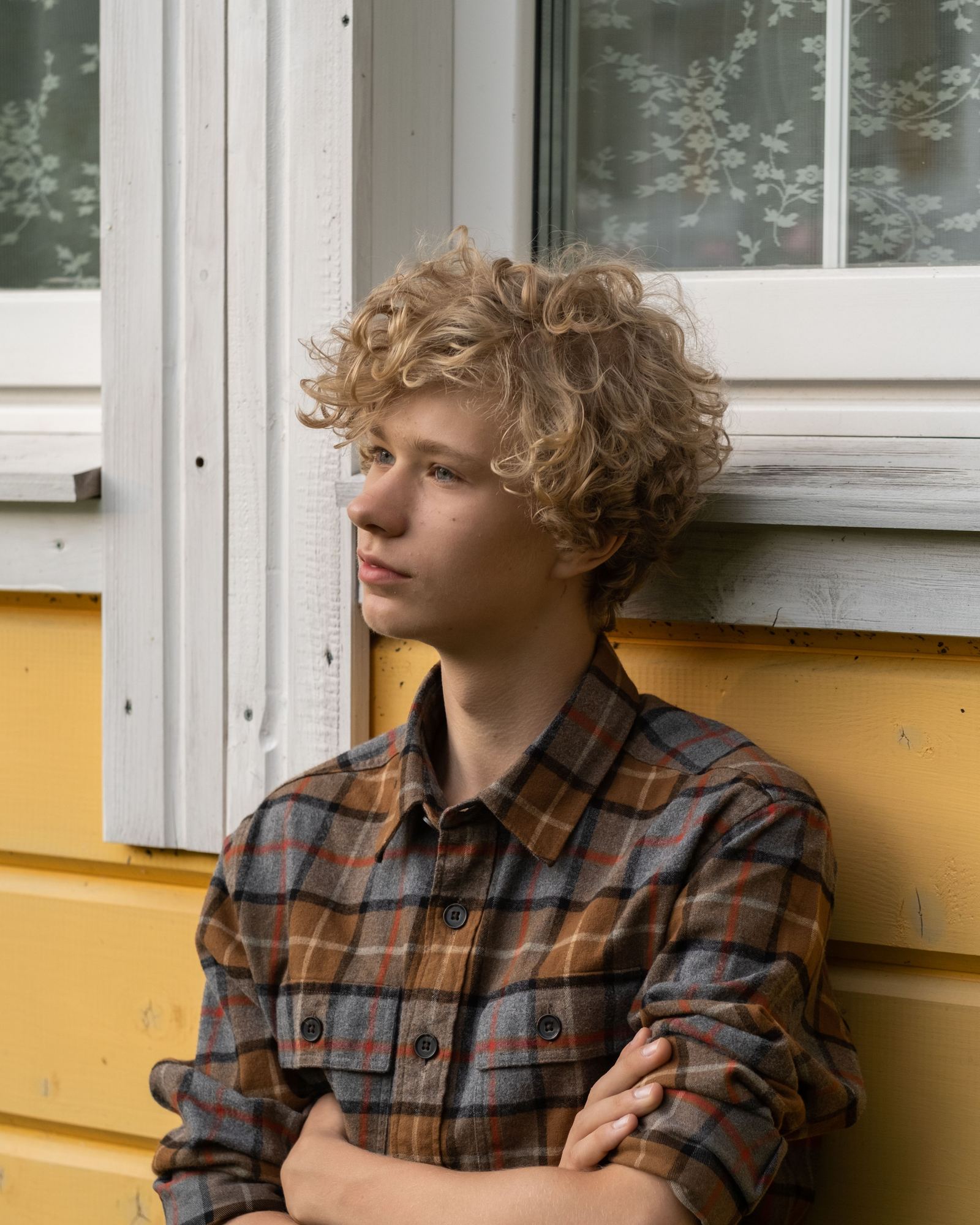 A teenager with curly hair standing by a window, arms crossed