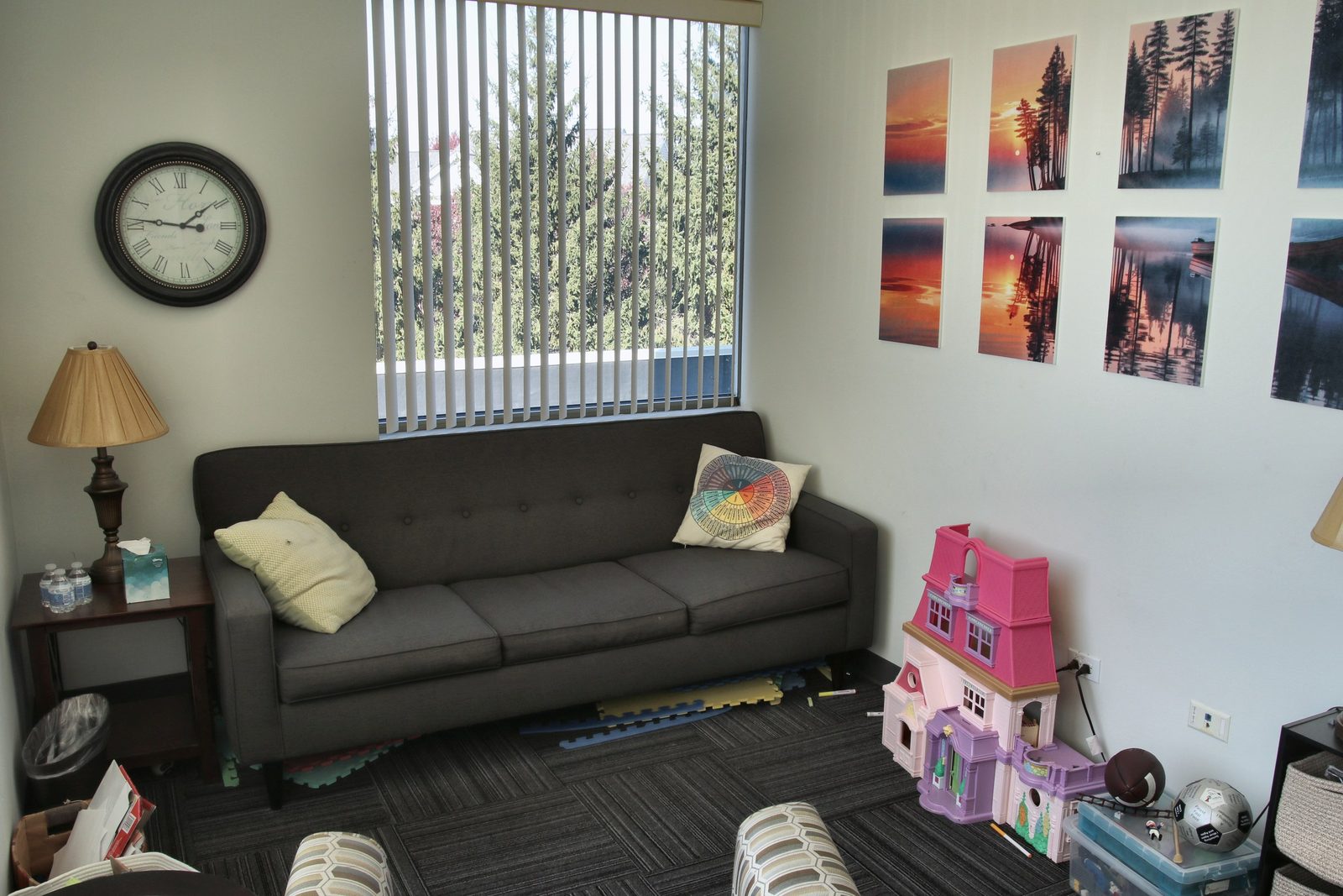 A counseling room at the Geneva office, with a dark sofa and bright pillows, a wall clock, colorful framed art, and a window with blinds.