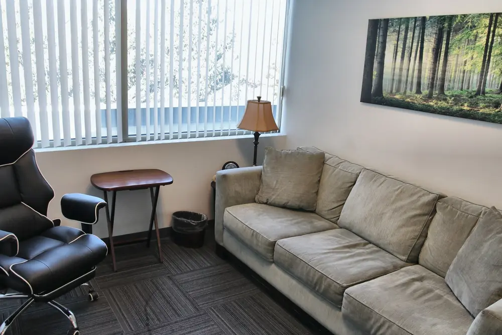 A counseling room at the Geneva office, with a beige sofa, a black desk chair, a floor lamp, and a forest print above a window with blinds.
