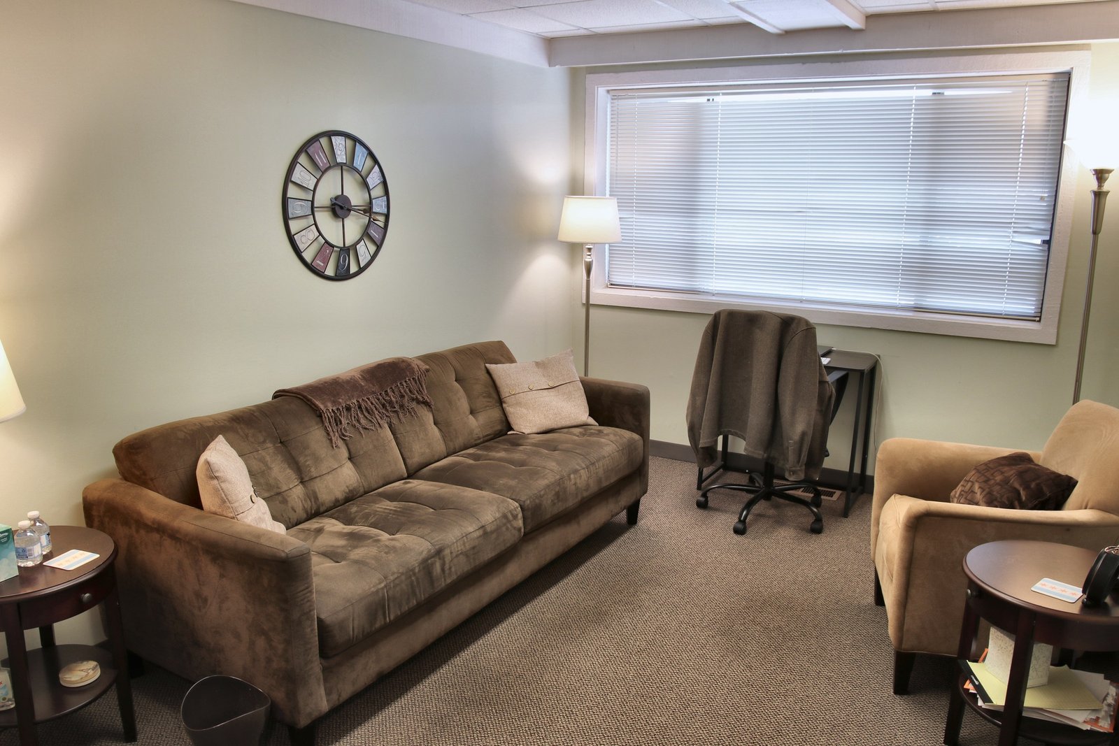 A counseling room at the Naperville office, with a brown sofa, a tan armchair, a large wall clock, and a window with blinds.