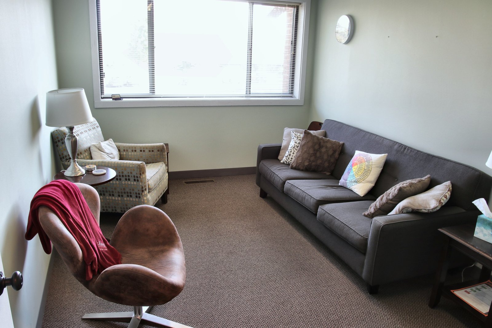 A counseling room at the Naperville office, with a gray sofa and pillows, a brown swivel chair with a red throw, and a patterned armchair by the window.