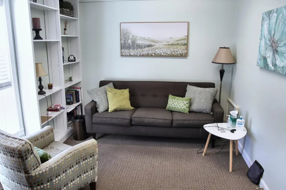 A counseling room at the Wheaton office, with a gray sofa and green pillows, a patterned armchair, a white bookshelf, and a landscape painting.