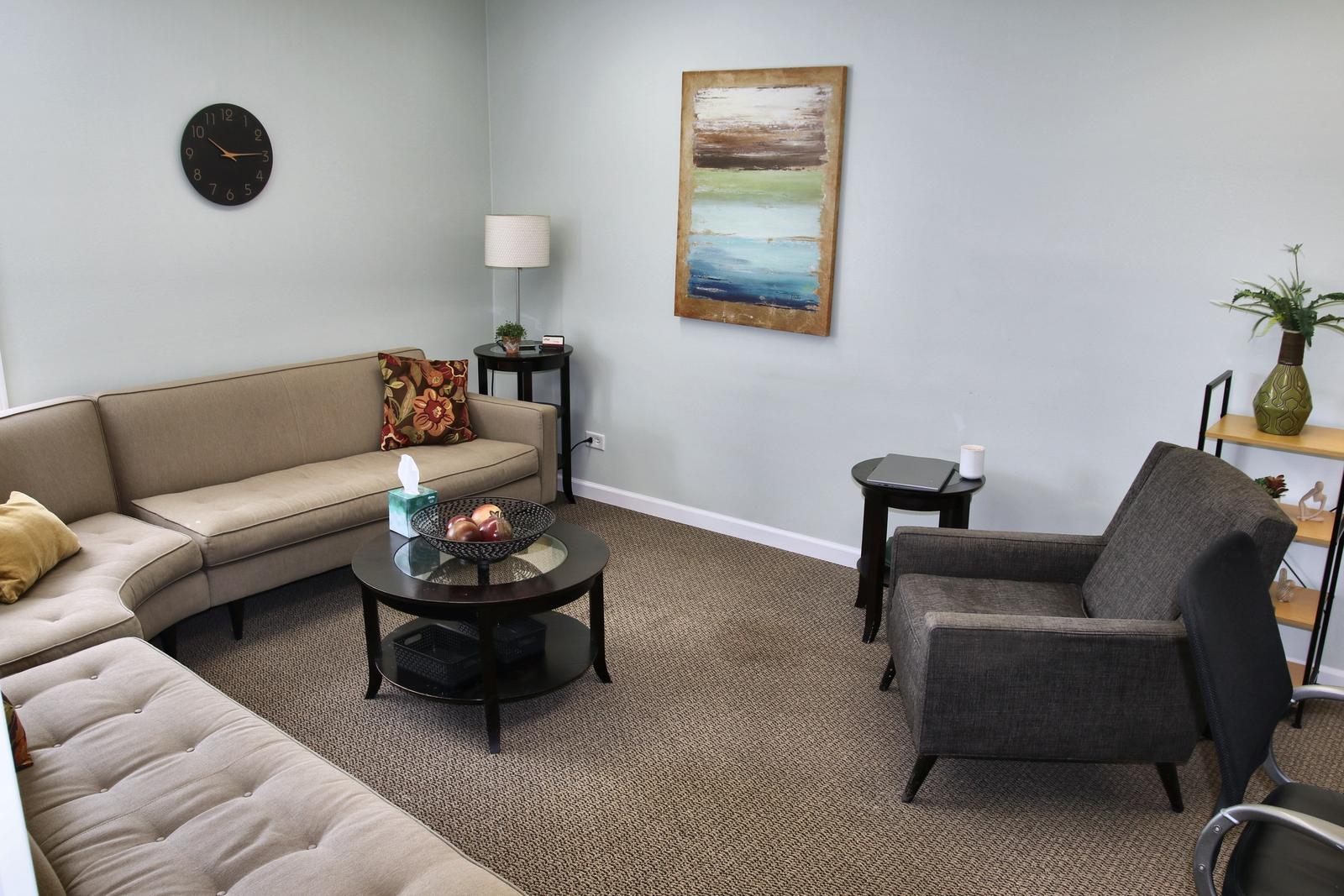 A counseling room at the Wheaton office, with a curved beige sectional, a round glass coffee table, a gray armchair, and an abstract painting.
