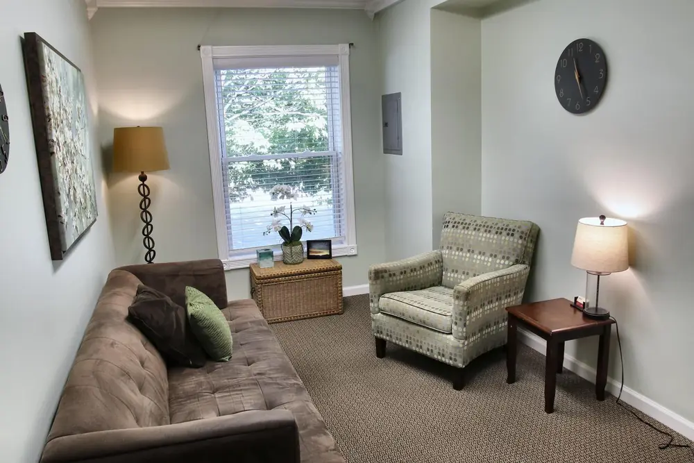 A counseling room at the Wheaton office, with a brown sofa, a patterned armchair, a lamp on a side table, and a window looking out on trees.