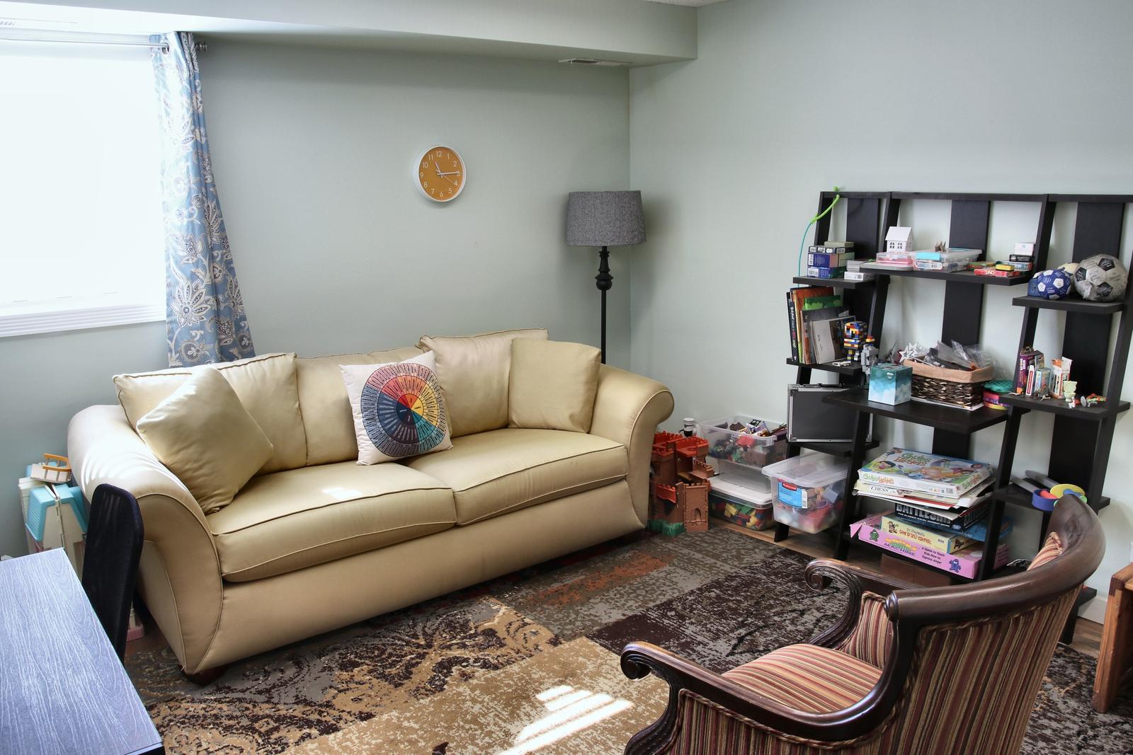 A play therapy room at the Wheaton office, with a tan sofa, a shelf of toys and games, and a patterned rug.