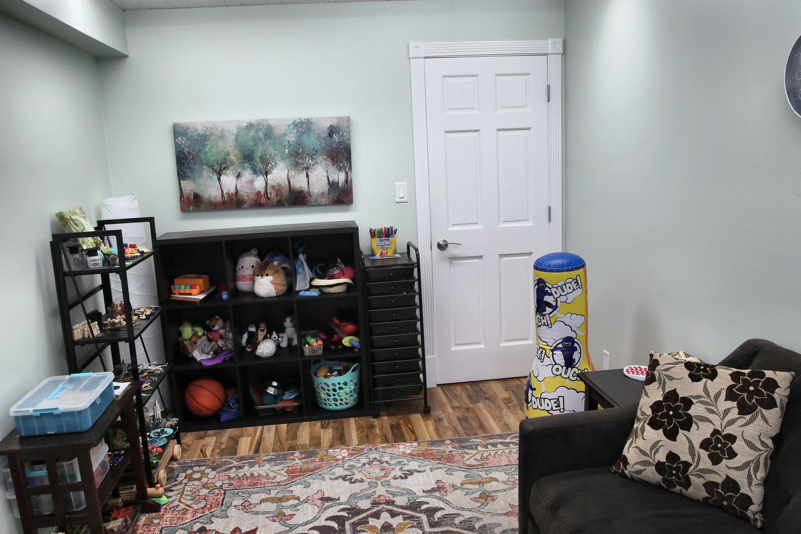 A play therapy room at the Wheaton office, with shelves of toys and art supplies, a floral pillow, and a painting of trees.