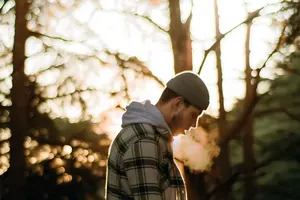 A man in a cap breathing out into cool morning air among the trees.