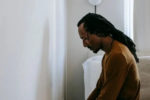 A man sitting alone on the edge of his bed, head bowed.