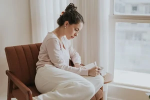 A young woman sitting calmly in a chair by a window, reading.